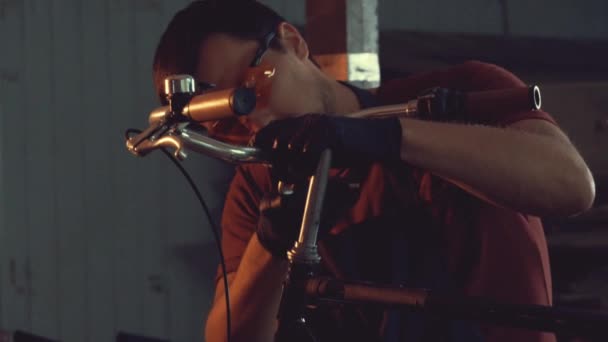 Tema pequeña empresa de reparación de bicicletas. joven caucásico morena hombre usando gafas de seguridad, guantes y delantal utiliza herramienta de mano reparación y ajuste de tallo y manillar de bicicleta en garaje del taller — Vídeo de stock