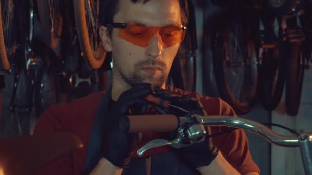 テーマ中小企業の自転車の修理。安全を身に着けているブルネット白人の若い男のゴーグル、手袋、エプロン、手のひらツールを使用して修復をワーク ショップのガレージに Handbrabar ブレーキ自転車のハンドルを調整します。 — ストック動画