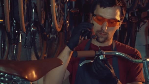 Tema pequeña empresa de reparación de bicicletas. Un joven caucásico morena hombre con gafas de seguridad, guantes y delantal utiliza herramienta de mano para reparar y ajustar el mango de la bicicleta de freno Handbrabar en el garaje del taller — Vídeos de Stock