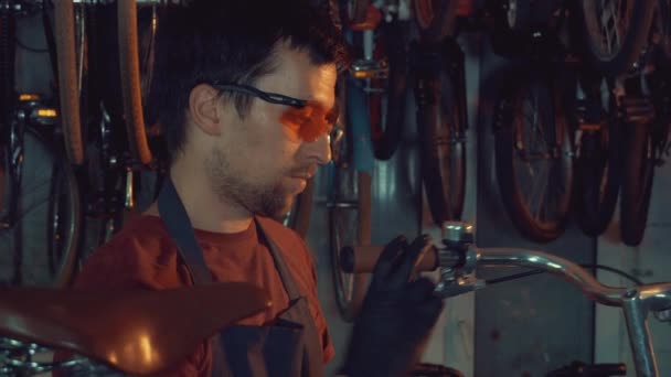 Thème petite entreprise réparation de vélo. Un jeune homme brune caucasien portant des lunettes de sécurité, des gants et un tablier utilise un outil à main pour réparer et ajuster la poignée de vélo de frein Handbrabar dans le garage de l'atelier — Video