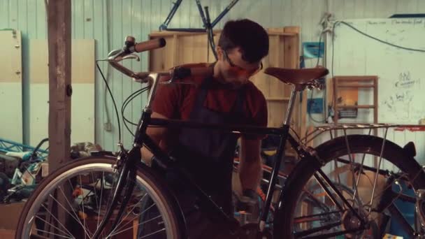 Tema småföretag cykelreparation. En ung brunett kaukasiska-man som bär skyddsglasögon, handskar och ett förkläde insisterar hastigheten växlar på cykel styret i garaget i verkstaden — Stockvideo