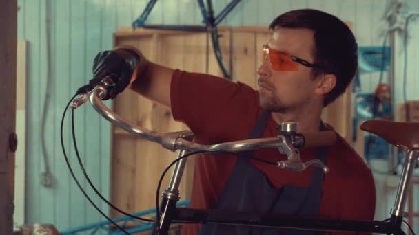 Thème petite entreprise réparation de vélo. Un jeune homme brune caucasien portant des lunettes de sécurité, des gants et un tablier insiste sur les interrupteurs de vitesse sur le guidon du vélo dans le garage de l'atelier — Video