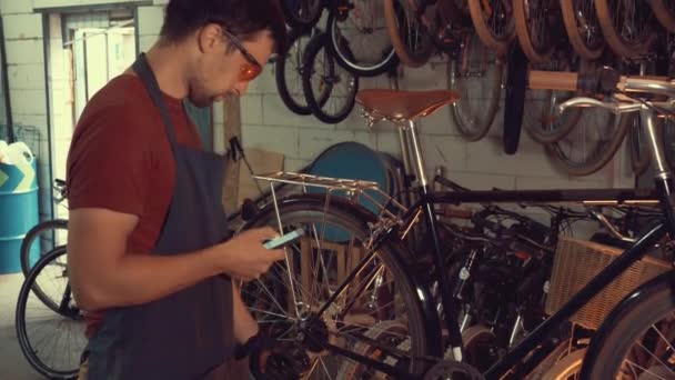 Téma kisvállalkozások kerékpár javítás. Kaukázusi barna fiatalember visel, védőszemüveg, kesztyű és fartukhe mobiltelefon technológiát használ, figyelembe veszi, a Feladatlista kerékpár műhely — Stock videók