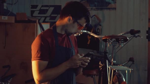 Theme small business bike repair. Young caucasian brunette man wearing safety goggles, gloves and fartukhe uses mobile phone technology, takes notes, checklist in bicycle workshop — Stock Video