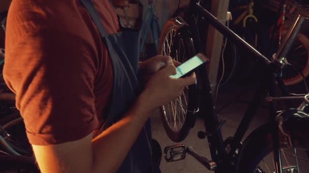 Tema pequeña empresa de reparación de bicicletas. Joven caucásico morena hombre con gafas de seguridad, guantes y fartukhe utiliza la tecnología del teléfono móvil, toma notas, lista de verificación en el taller de bicicletas — Vídeo de stock