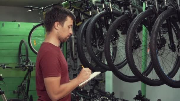 Thema Kleinunternehmen, die Fahrräder verkaufen. junge kaukasische männliche Brünette Kleinunternehmer, Filialleiter verwendet Notizblock und Stift macht Notizen, Checkliste im Fahrradgeschäft — Stockvideo