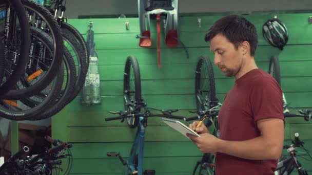 Theme of small business selling bicycles. Young Caucasian male brunette small business owner, store manager uses notepad and pen makes notes, checklist at bicycle store — Stock Video
