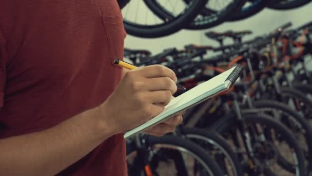 Tema de la pequeña empresa venta de bicicletas. Joven caucásico hombre morena dueño de una pequeña empresa, gerente de la tienda utiliza bloc de notas y bolígrafo toma notas, lista de verificación en la tienda de bicicletas — Vídeo de stock