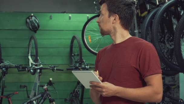 Tema for småhandel med sykler. Ung, hvit, mannlig brunette, liten bedriftseier, butikksjef bruker notisblokk og penn, sjekkliste i sykkelbutikk – stockvideo