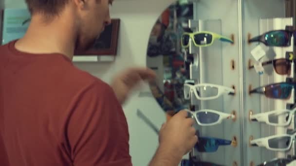 Um jovem caucasiano está de pé perto de um corrimão óculos esportivos para esportes, ciclismo e escolhe óculos em uma loja — Vídeo de Stock