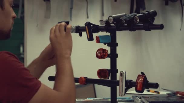 A young Caucasian man stands near the shelter and chooses, presses the button and turns off the bicycle flashing back light for a safe ride on the road at the bicycle store — Stock Video