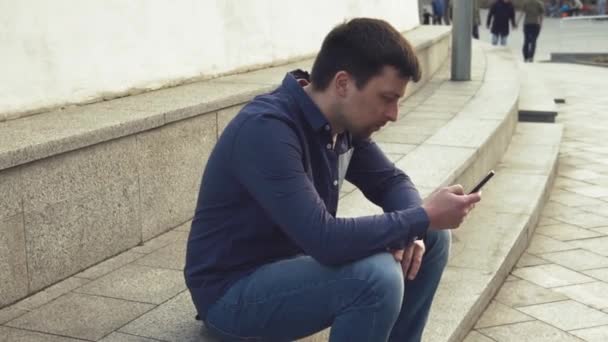 Tema es el hombre y las redes sociales. joven caucásico triste hombre morena en camisa está sentado en el parque en Letnec esperando, llegar tarde está utilizando la mano para sostener el teléfono móvil. Emoción resentimiento y trastorno — Vídeos de Stock