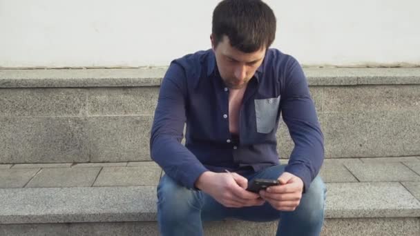 Tema es el hombre y las redes sociales. joven caucásico triste hombre morena en camisa está sentado en el parque en Letnec esperando, llegar tarde está utilizando la mano para sostener el teléfono móvil. Emoción resentimiento y trastorno — Vídeos de Stock