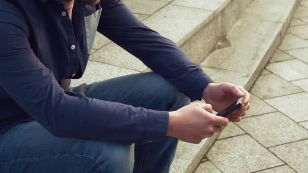 Tema è l'uomo e le reti sociali. giovane caucasico triste bruna maschio in camicia è seduto nel parco in attesa Letnec, essendo in ritardo sta usando la mano per tenere il telefono cellulare. Emozione risentimento e disordine — Video Stock