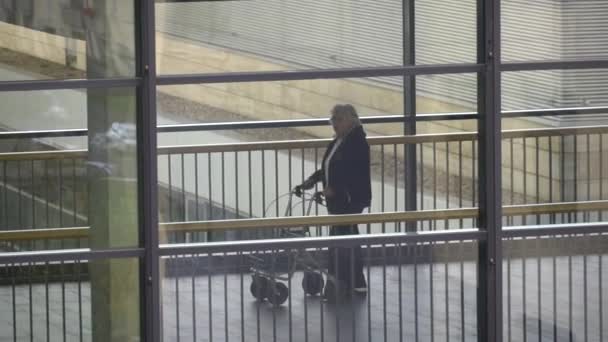 20. oktober 2018. krefeld deutschland. Thema Medizin und Gesundheit. alte reife kaukasische Patientin in der Klinik geht mit Rollator drinnen entlang der Brücke zwischen den Stationen im Flur. Blick aus dem Fenster — Stockvideo