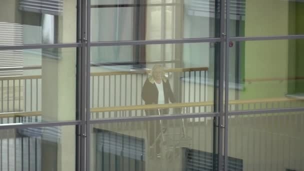 20 жовтня 2018. Крефельд. Тема медицини та охорони здоров'я. Старий зрілих кавказьких пацієнтки в клініці прогулянки з Уокер всередині вздовж мосту між станціями в коридорі. Переглянути через вікно — стокове відео