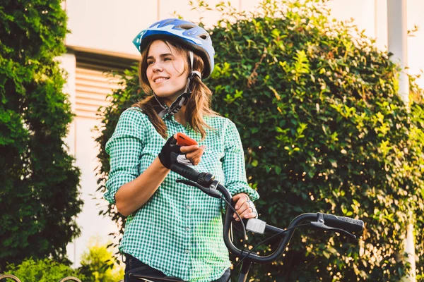 Genç beyaz kız öğrenci el holding telefon dokunmatik ekran için kullanır. Bir kadın bir kiralama şehir bisikleti bir kask ve gömlek bir kaldırımda güneşli havalarda durumda. Mola mola — Stok fotoğraf