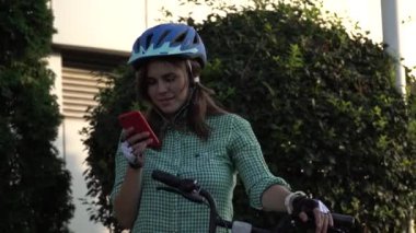 Genç beyaz kız öğrenci el holding telefon dokunmatik ekran için kullanır. Bir kadın bir kiralama şehir bisikleti bir kask ve gömlek bir kaldırımda güneşli havalarda durumda. Mola mola
