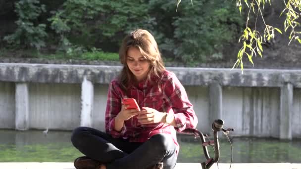 Thema ökologischer Fahrradtransport. junge kaukasische Frau im Hemd Studentin sitzt in einem Park in der Nähe des Sees zum Ausleihen orangefarbenes Fahrrad verwendet ein Handy Herbstferien bei sonnigem Wetter Technologie — Stockvideo