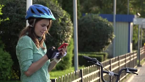Young caucasian female student uses hand holding phone to touch screen. A woman stands near a rental city bike in sunny weather on a sidewalk in a helmet and shirt. Rest stop break — Stock Video