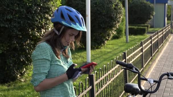 Joven estudiante caucásica utiliza la mano sosteniendo el teléfono para tocar la pantalla. Una mujer se para cerca de una bicicleta de alquiler de la ciudad en un clima soleado en una acera con un casco y una camisa. Descanso stop break — Vídeo de stock