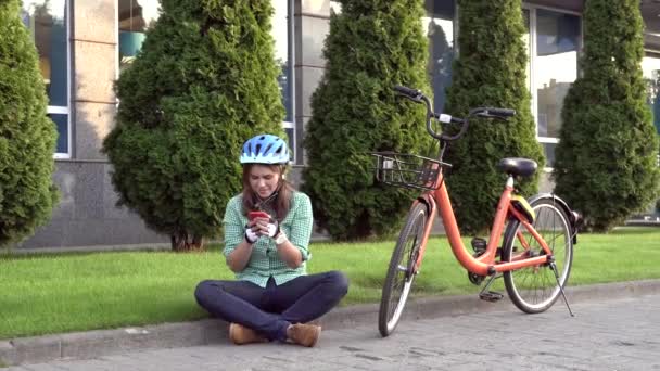 Sujeito modo ecológico de transporte de bicicleta. Mulher branca nova bonita vestindo um capacete azul e poses longas do cabelo que estão ao lado de uma bicicleta alaranjada do aluguer com uma cesta em um parque da cidade — Vídeo de Stock