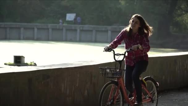 Asunto transporte ecológico de bicicletas. Joven mujer caucásica montando en un camino de tierra en un parque cerca de un lago alquilando una bicicleta de color naranja en otoño en un clima soleado — Vídeos de Stock