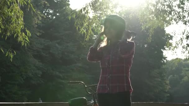 Soggetto ecologico modo di trasporto bicicletta. Bella giovane donna kasazy che indossa un casco blu e pose di capelli lunghi in piedi accanto a una bici a noleggio di colore arancione con un cesto in un parco cittadino — Video Stock