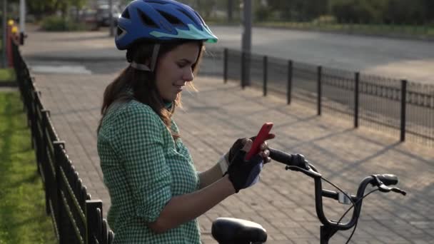 Joven estudiante caucásica utiliza la mano sosteniendo el teléfono para tocar la pantalla. Una mujer se para cerca de una bicicleta de alquiler de la ciudad en un clima soleado en una acera con un casco y una camisa. Descanso stop break — Vídeos de Stock