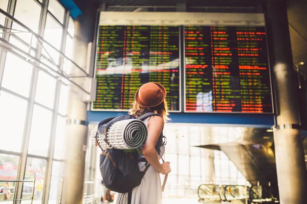 Tema viajes transporte público. mujer joven de pie con la espalda en vestido y sombrero detrás de la mochila y el equipo de camping para dormir, alfombra aislante se ve horario en la estación del aeropuerto marcador — Foto de Stock