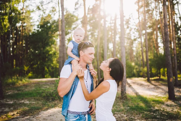 Família mãe, pai e filha se senta no papai nos ombros, e os pais se beijam na natureza na floresta no verão ao pôr do sol — Fotografia de Stock