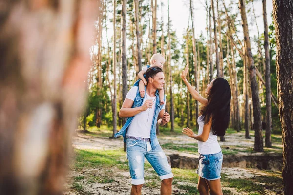 Een familie van drie, jonge mooie ouders spelen met hun dochter zit op de schouders van de paus een jaar na de geboorte in de naaldhout bos in de zomer — Stockfoto