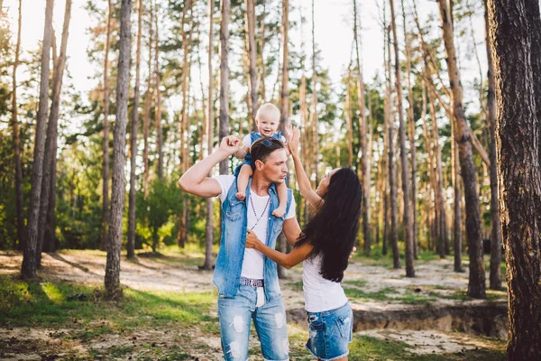 a family of three, young beautiful parents playing with their daughter sitting on the shoulders of the pope one year after birth in the coniferous forest in summer