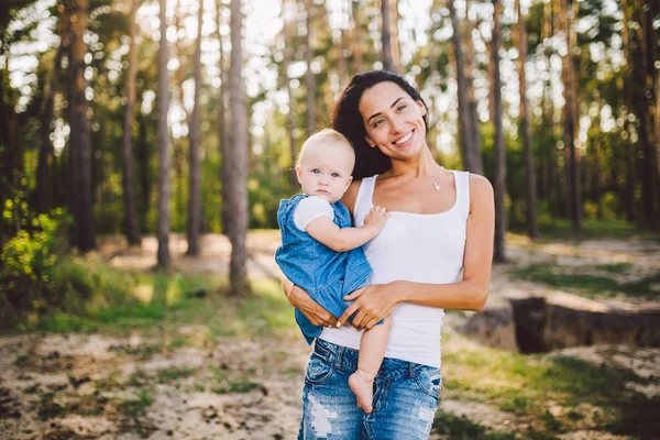 young beautiful fashionable mother with long brunette hair holding a daughters blonde with blue eyes one year of birth in a coniferous forest in a summer park.