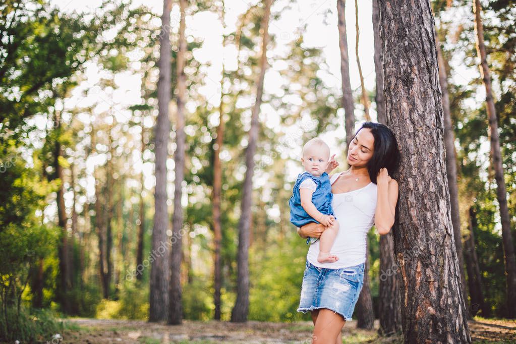 young beautiful fashionable mother with long brunette hair holding a daughter's blonde with blue eyes one year of birth in a coniferous forest in a summer park. Wearing jeans clothes
