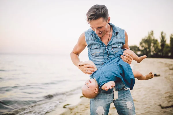 Thema vaderschap en de rest met een kind op zee. Jonge knappe Kaukasische vader speelt speelt geniet overgeven omhoog in wapens zijn kind in zomer dochter op het zandstrand aan de kust — Stockfoto