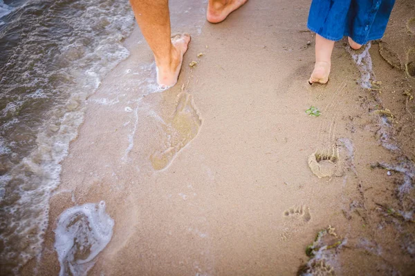 Joven padre caucásico en pantalones vaqueros descalzo va y sostiene la mano de una hija de un año con la espalda a lo largo de la playa cerca del mar. Papá le enseña a caminar al niño. Huellas en la arena — Foto de Stock