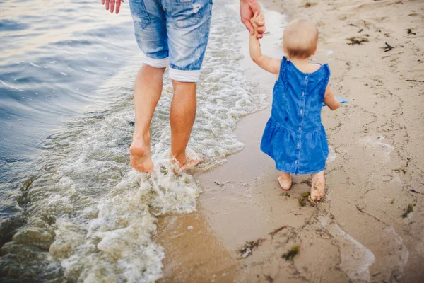 Joven padre caucásico en pantalones vaqueros descalzo va y sostiene la mano de una hija de un año con la espalda a lo largo de la playa cerca del mar. Papá le enseña a caminar al niño. Huellas en la arena —  Fotos de Stock