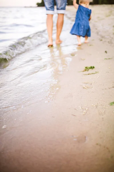 Joven padre caucásico en pantalones vaqueros descalzo va y sostiene la mano de una hija de un año con la espalda a lo largo de la playa cerca del mar. Papá le enseña a caminar al niño. Huellas en la arena —  Fotos de Stock