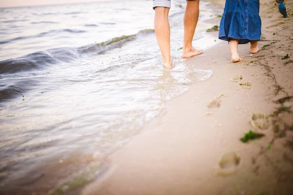 Joven padre caucásico en pantalones vaqueros descalzo va y sostiene la mano de una hija de un año con la espalda a lo largo de la playa cerca del mar. Papá le enseña a caminar al niño. Huellas en la arena —  Fotos de Stock