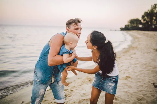 En ung, vacker familj på tre. Mamma, Pappa och dotter i armarna på min far spela, jubla, leende på sandstranden på stranden på sommaren. Klädd i junky kläder och vita skjortor — Stockfoto