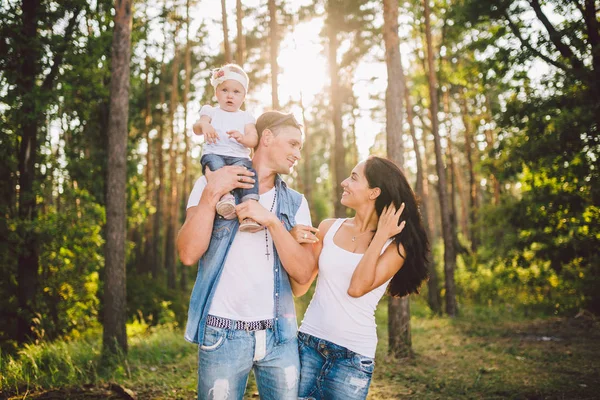 Stilren ung familj mamma, pappa och dotter ett år gammal blondin sitter nära far på axlar, utomhus utanför staden i en park mitt bland träd på sommaren. Bär jeans kläder. Familjefotografering — Stockfoto