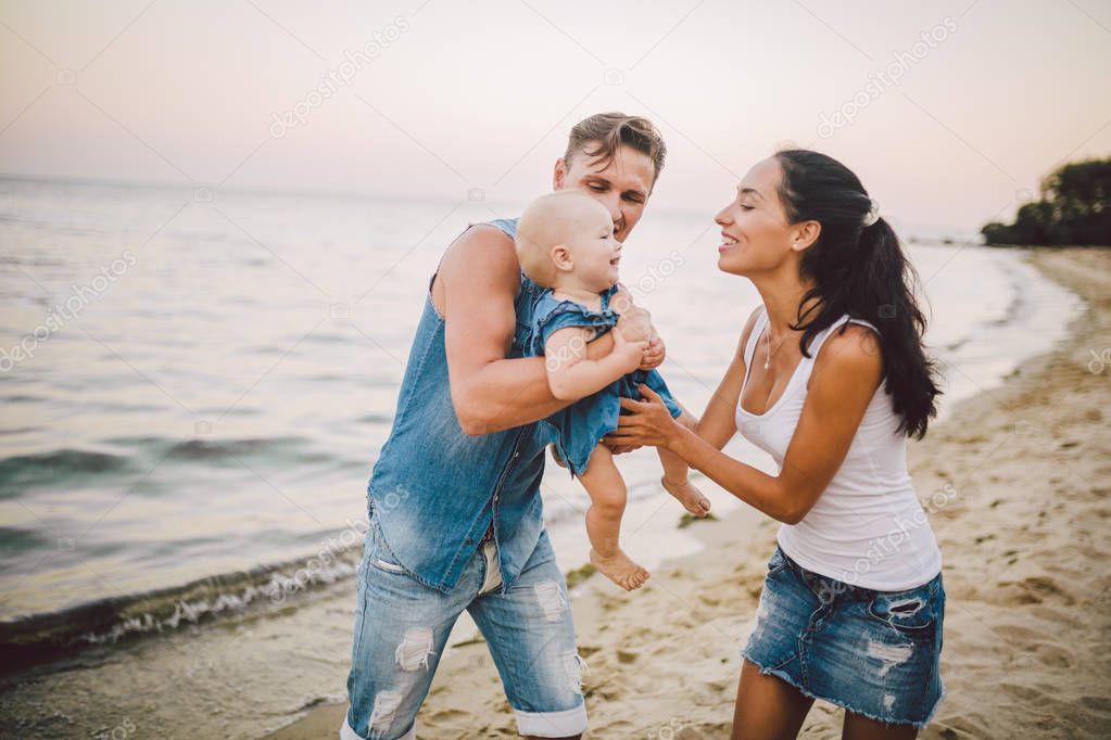 A young, beautiful family of three. Mom, Dad and daughter in the arms of my father play, rejoice, smile on the sandy beach on the beach in the summer. Dressed in junky clothes and white shirts
