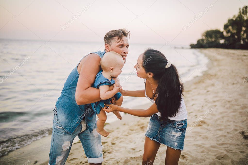 A young, beautiful family of three. Mom, Dad and daughter in the arms of my father play, rejoice, smile on the sandy beach on the beach in the summer. Dressed in junky clothes and white shirts