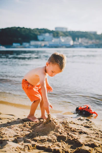 Sujeto construcción e industria pesada. Abstracción niño jugando en la arena cerca del río en el modelo de tractor rojo de juguete de verano, máquina excavadora con un cubo —  Fotos de Stock