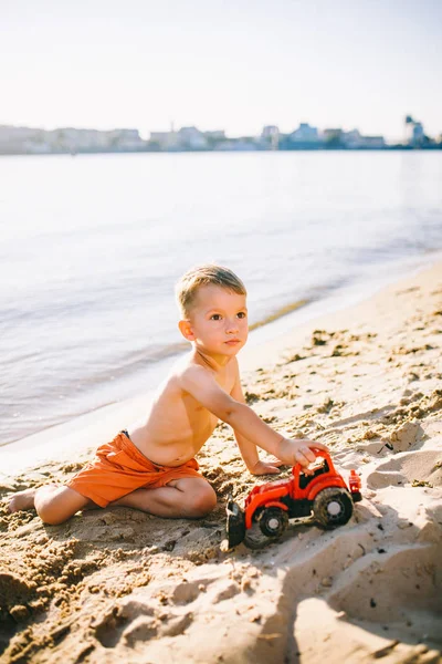 Sujeto construcción e industria pesada. Abstracción niño jugando en la arena cerca del río en el modelo de tractor rojo de juguete de verano, máquina excavadora con un cubo —  Fotos de Stock