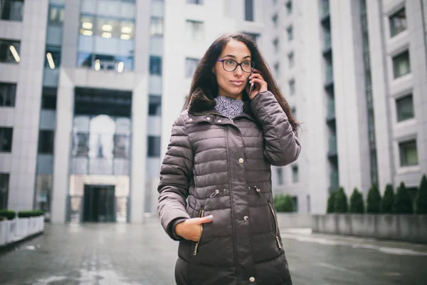 El tema es la situación del negocio. Hermosa mujer joven de etnia europea con el pelo largo y morena con gafas y percheros en el fondo del centro de negocios y utiliza el teléfono en la mano cerca de la oreja — Foto de Stock