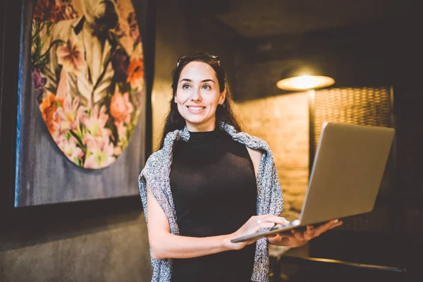 Het concept van kleine business en technologie. Jonge mooie brunette zakenvrouw in zwarte jurk en grijze trui staat in de koffieshop met laptop in handen. Op het hoofd bril voor zicht — Stockfoto