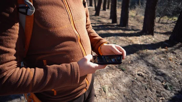 Theme tourism and technology. Young caucasian man with beard and backpack. Hiking tourist in pine forest uses technology, hand holding mobile phone to touch the screen. Gps application orientation — Stock Photo, Image