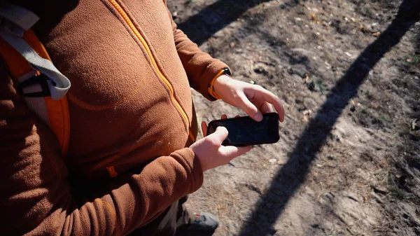 Theme tourism and technology. Young caucasian man with beard and backpack. Hiking tourist in pine forest uses technology, hand holding mobile phone to touch the screen. Gps application orientation — Stock Photo, Image